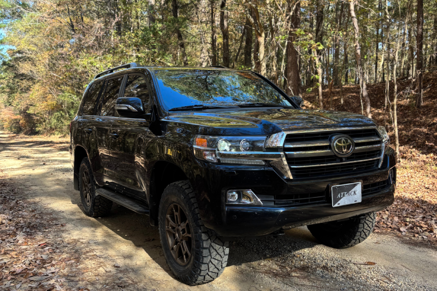 A black Toyota Land Cruiser 200 Series parked on a dirt trail surrounded by autumn trees, featuring bronze wheels and rugged off-road tires