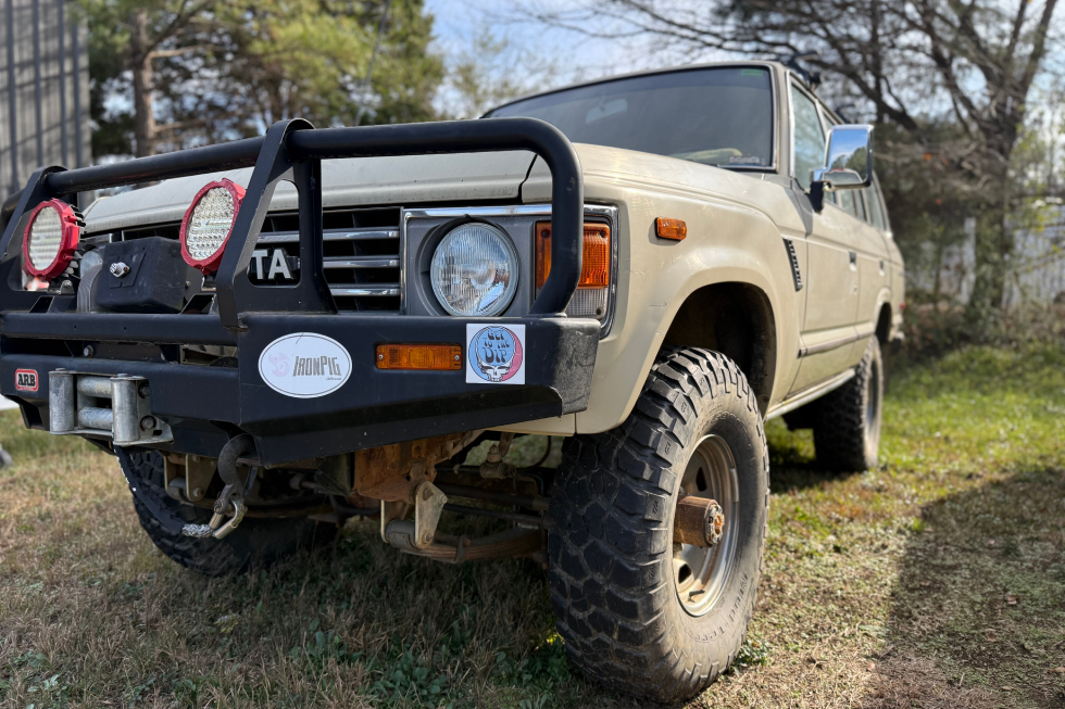 A tan Toyota Land Cruiser FJ60 equipped with an ARB bull bar bumper, off-road tires, and auxiliary lights, parked on grass