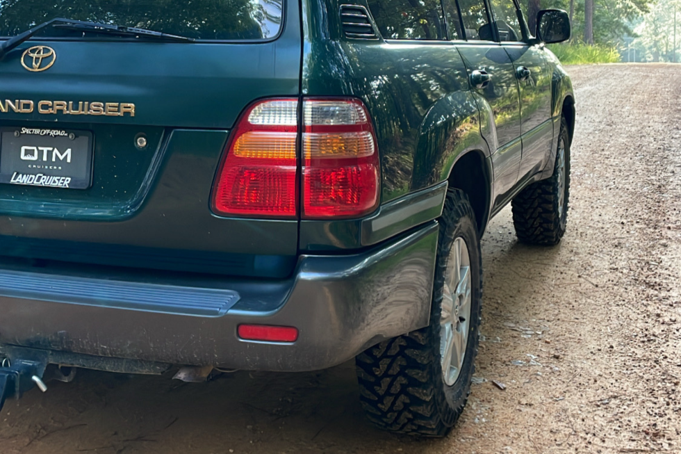 Rear-quarter view of a dark green Toyota Land Cruiser 100 Series trail truck fitted with rugged mud-terrain tires and an OTM Cruisers custom license plate frame, parked on a dirt path