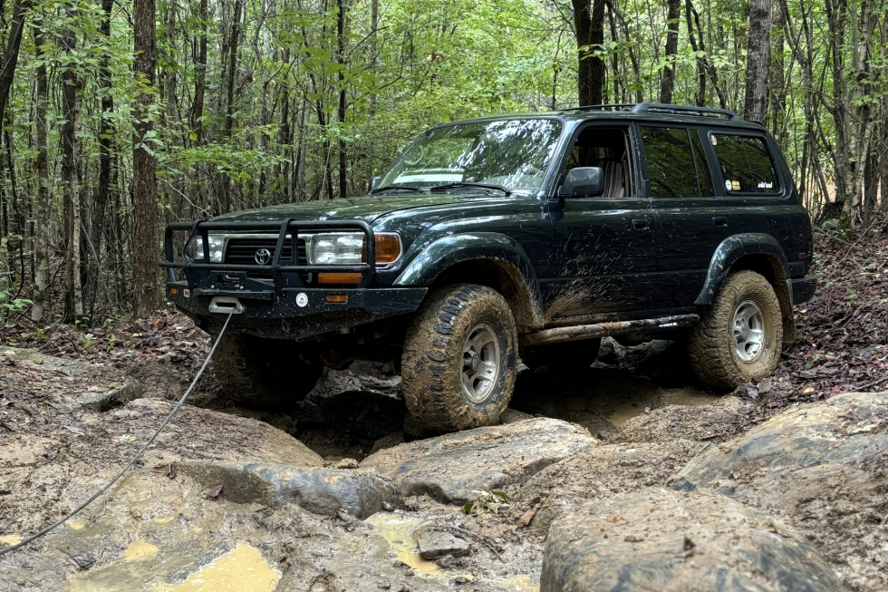 A dark green Toyota Land Cruiser 80 Series navigating a muddy, rocky off-road trail using its front bumper winch